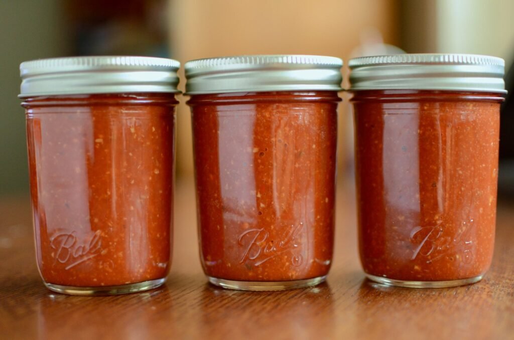 Three half pint jars of homemade harissa sauce, sitting in a row on a wooden table.